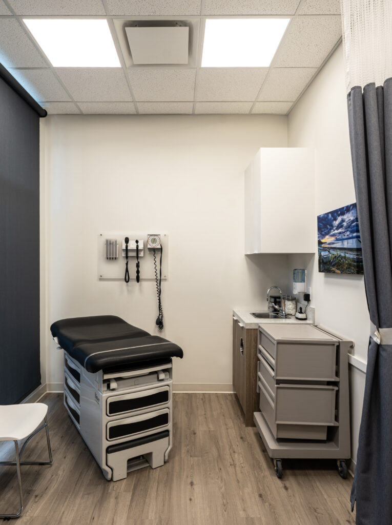 Medical exam room with an adjustable padded exam table, wall medical instruments, white cabinetry, and a sink with a counter on the right; gray curtains along the wall.