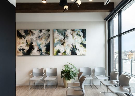 Modern interior showing white stacked chairs arranged in rows, abstract paintings on the wall, and large windows on the right with a potted plant nearby.
