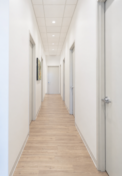 Bright white hallway with gray doors along both sides and a light wood floor, leading to a doorway in the distance.